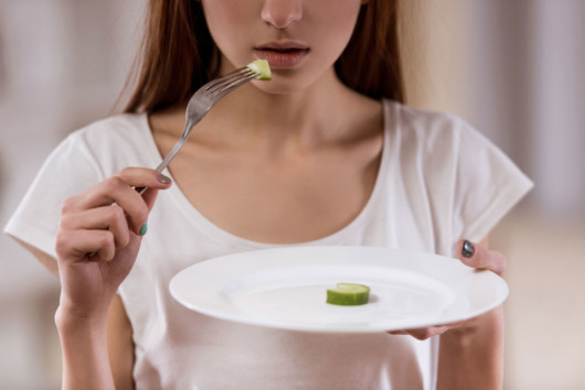 Young girl is holding a plate with one piece cucumber on it and a fork to her mouth