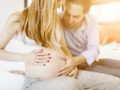 A young happy couple is sitting on the sofa next to each other. The man is touching his pregnant wife's belly