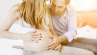 A young happy couple is sitting on the sofa next to each other. The man is touching his pregnant wife's belly