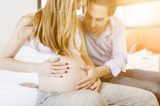 A young happy couple is sitting on the sofa next to each other. The man is touching his pregnant wife's belly