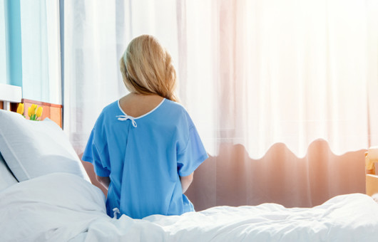 The girl is sitting in the hospital blue gown on the bed after the tummy tuck operation