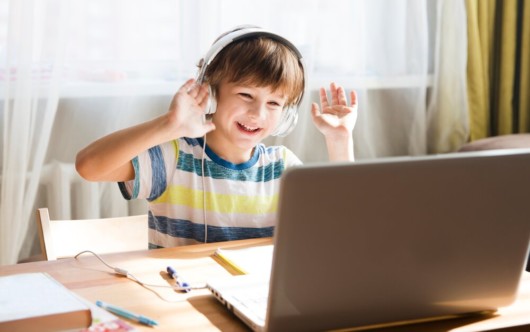 A boy is sitting in the headphoneswaving a hand in front of the laptop and having speech teletherapy