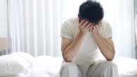 A young guys sits on the bed and covers his face with hands