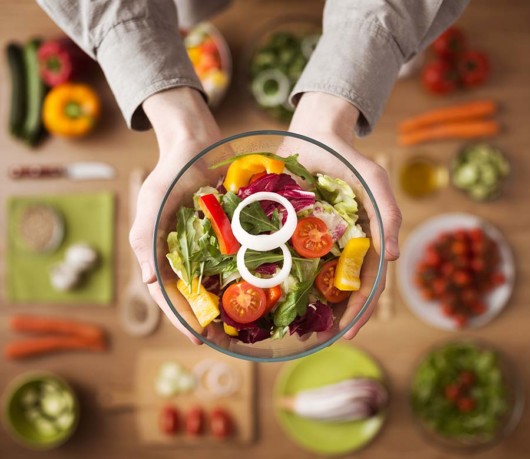 A man is holding a bowl of salad in his hands. At the background there are vegetables and fruits