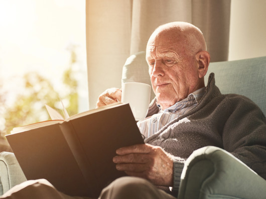 A senior man is reading a book and drinking tea in his chair