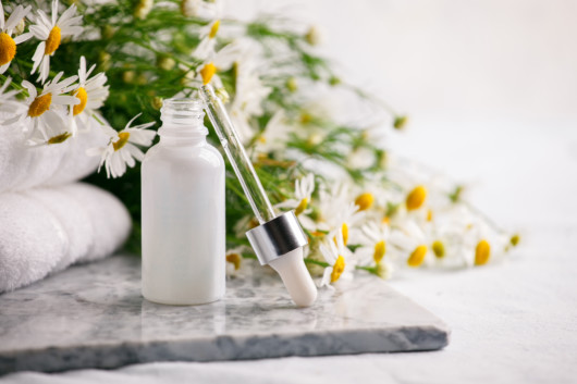 Essential camomile oil in white bottle next to the camomile flowers at the background