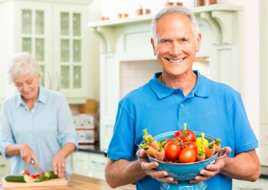 A senior man is holding a bowl of fresh veggies and smiling and the senior lady at the background is cooking