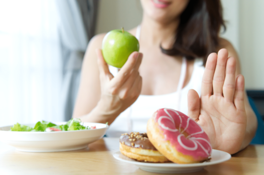 A girl is sitting at the table and holding a green apple in one hand and she rejects the junk food with the other one