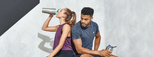A guy and the girl in sport outfit are sitting against grey wall. The girl is drinking pre-workout and the guy is holding the bottle and looking at her
