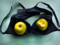 A black bra lying upside down on the green table and two green apples are inside the cups of the bra