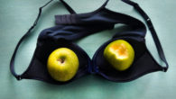 A black bra lying upside down on the green table and two green apples are inside the cups of the bra