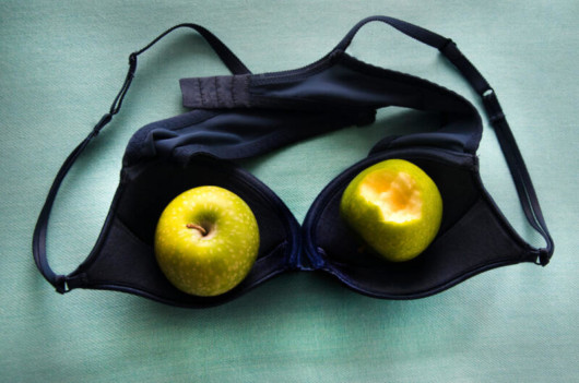 A black bra lying upside down on the green table and two green apples are inside the cups of the bra