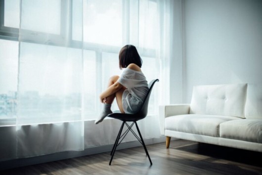 A young girl is sitting on the chair embracing her legs and looking into the window