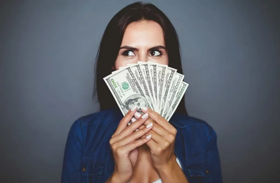 A woman on blue is holding a roll of dollars banknotes in her hands and in front of her face