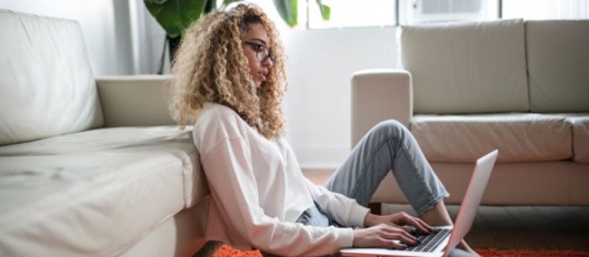 A young girl with curly hair is surfing the internet on the laptop sitting on the floor