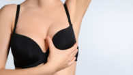 Young woman in black bra holding her breast on light background.
