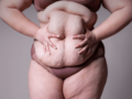 A woman with excessive weight in beige underwear on grey background holding her big belly