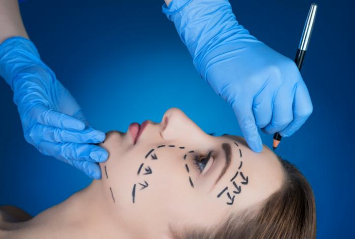 A doctor is putting marks on girl's face on blue background before the facelift procedure