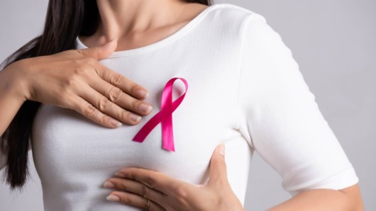 A close up picture of a woman in white top with a pinky cancer ribbon on her breast on white background who is holding her breast